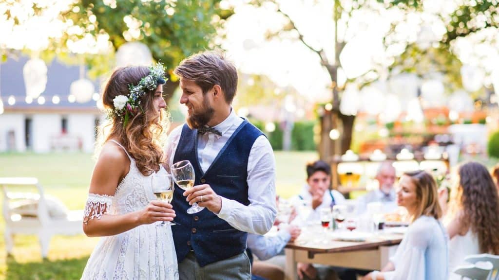 A bride wearing a flower crown and a groom in a vest clinking wine glasses outdoors.