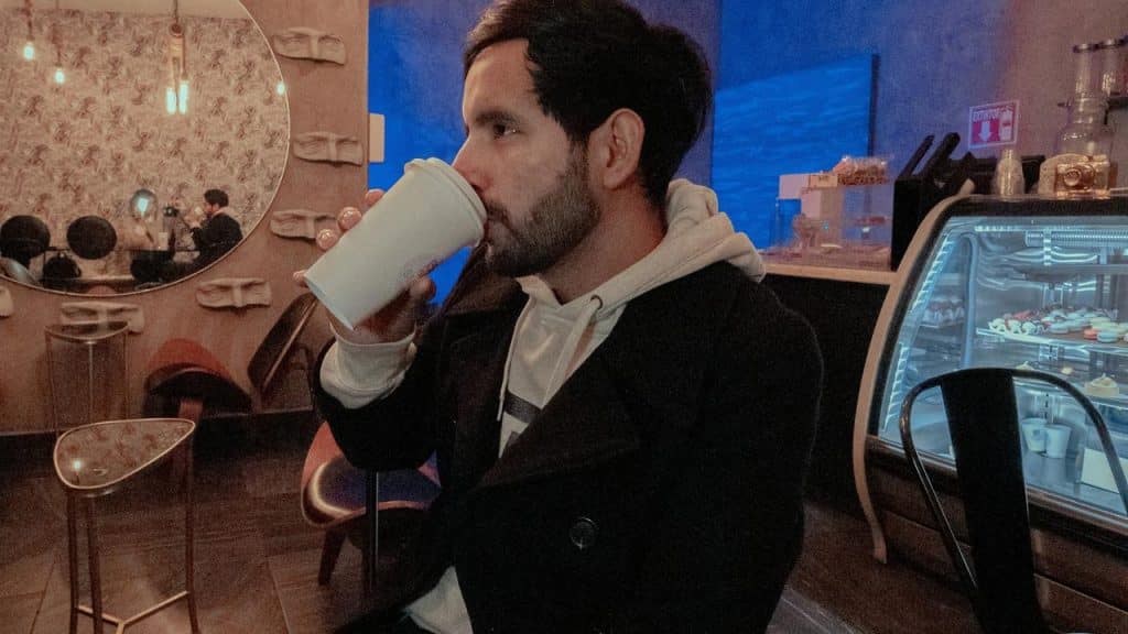 A man sipping coffee inside a softly lit café with a pastry display nearby.