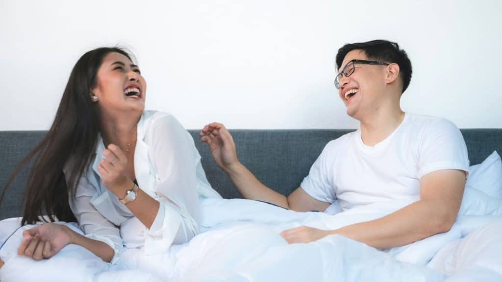 A man and woman sit in bed together, laughing with their heads tilted back.