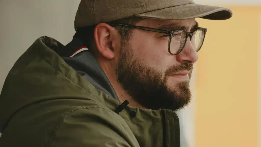 A bearded man wearing glasses and a cap, looking to the side.