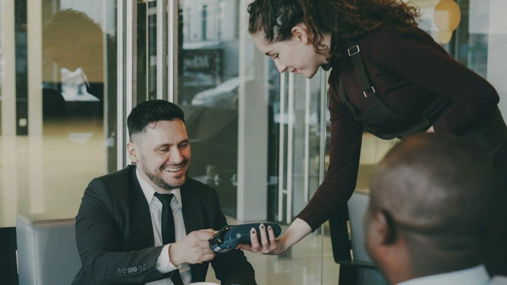 A man in a suit smiles while tapping a card on a payment terminal held by a server.