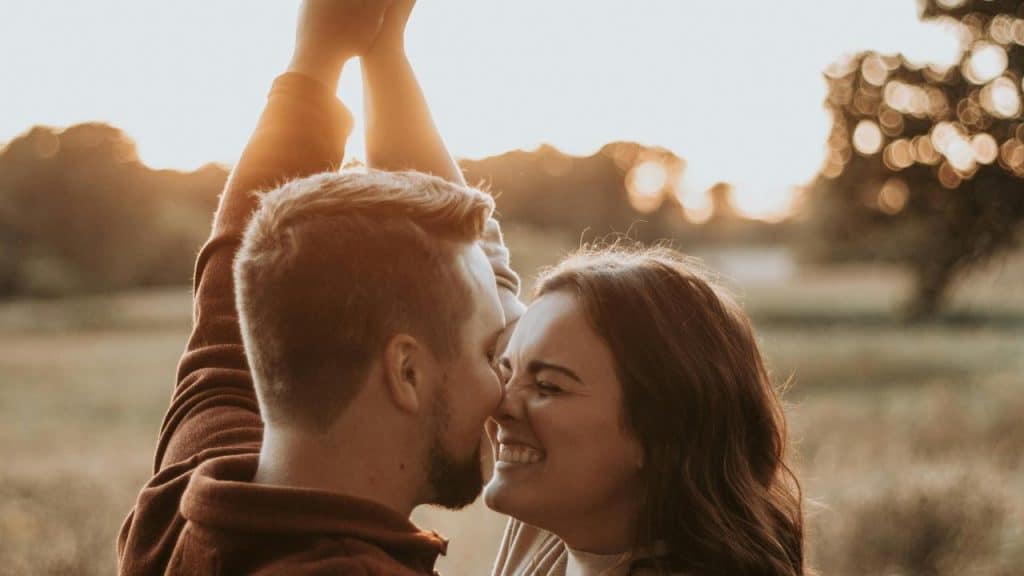 A couple smiling and touching noses while raising their arms together at sunset.