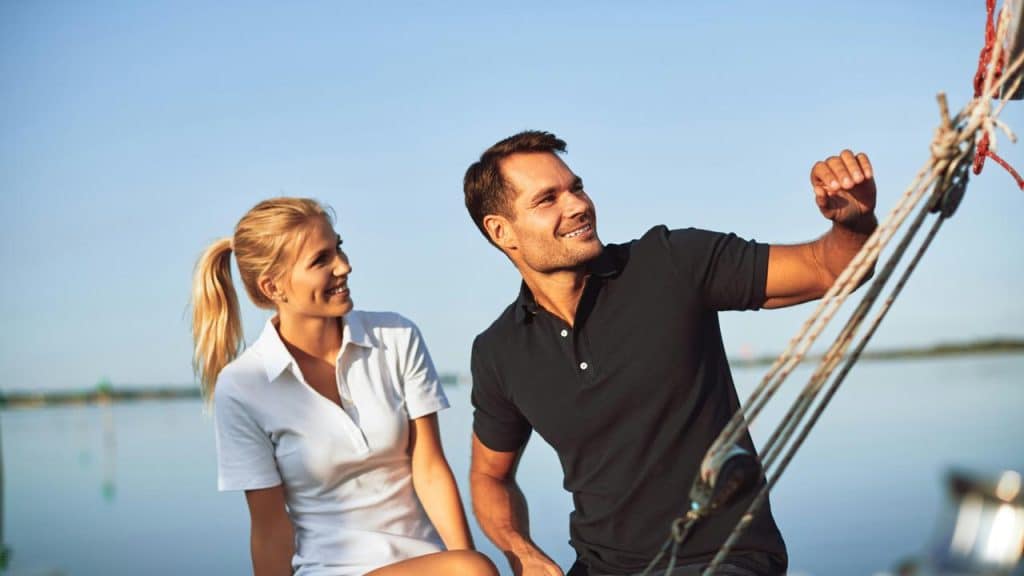 A smiling couple on a sailboat adjusting rigging on calm water.