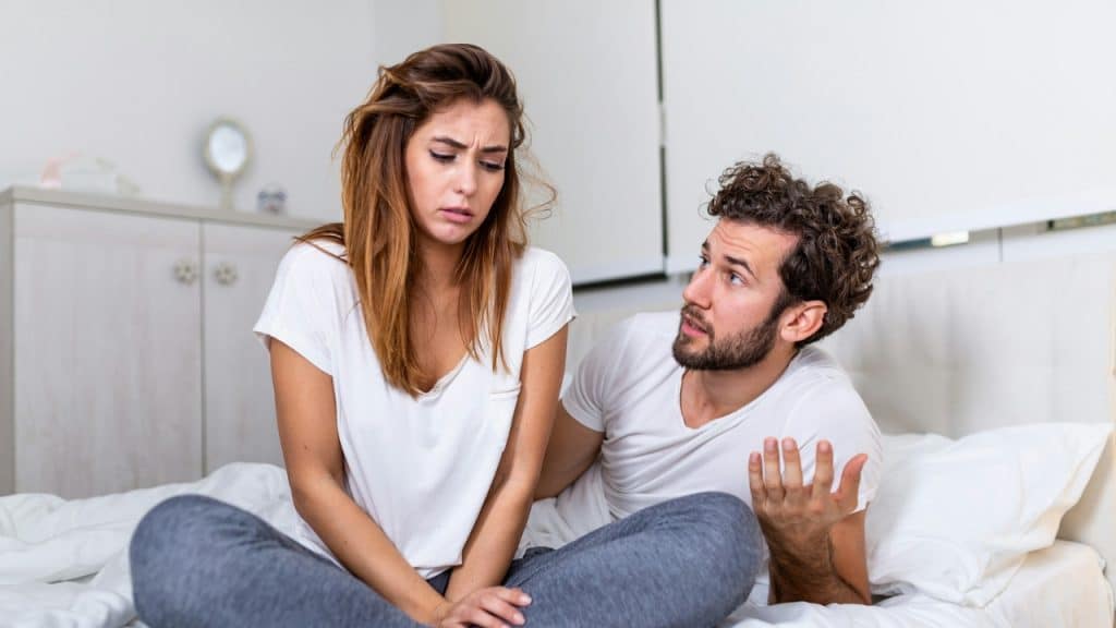 A man gestures with one hand while talking to a woman sitting on a bed.