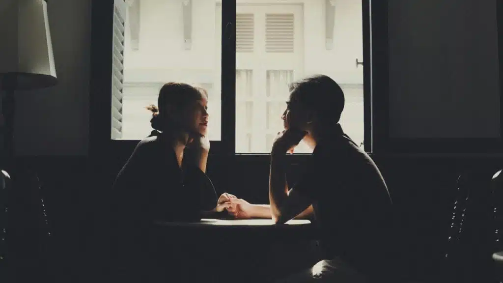 A man and woman sit in silhouette at a table, facing each other before a window.