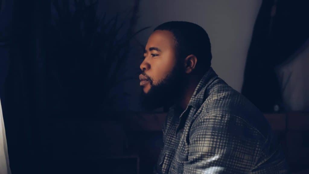 A bearded man in a plaid shirt sits in a dark room looking toward a window.