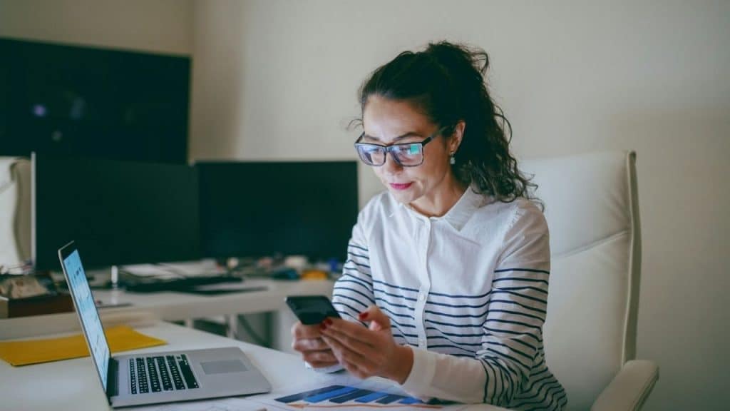 A woman in a striped shirt and glasses sits at a desk using her smartphone.