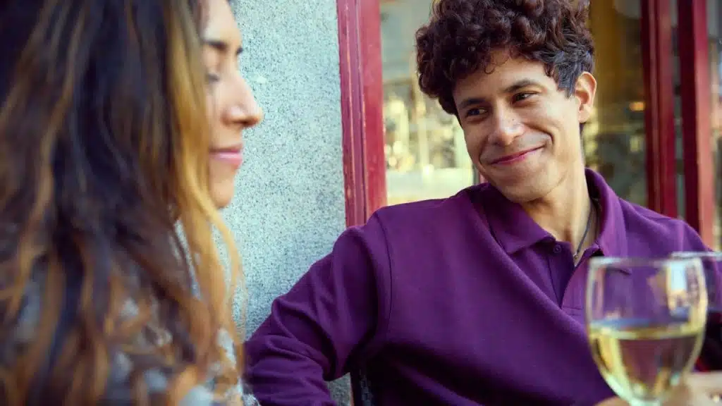 A man with curly hair smiles while looking at a woman during a conversation outdoors.