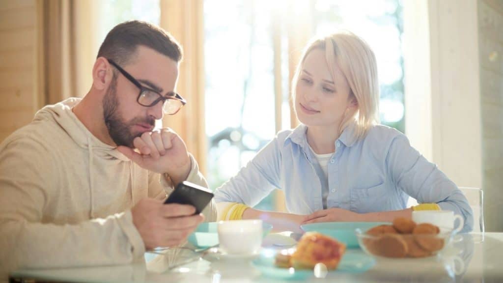 A man with glasses looks at his phone while a woman watches him across a table.