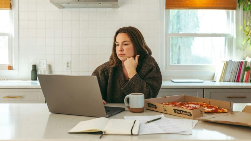 A woman looks at a laptop beside a pizza box and coffee mug in a kitchen.
