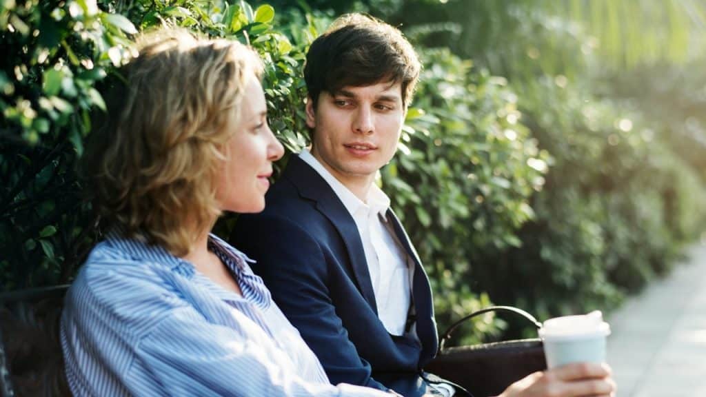 A man in a suit jacket looks toward a woman in a striped shirt sitting outdoors.