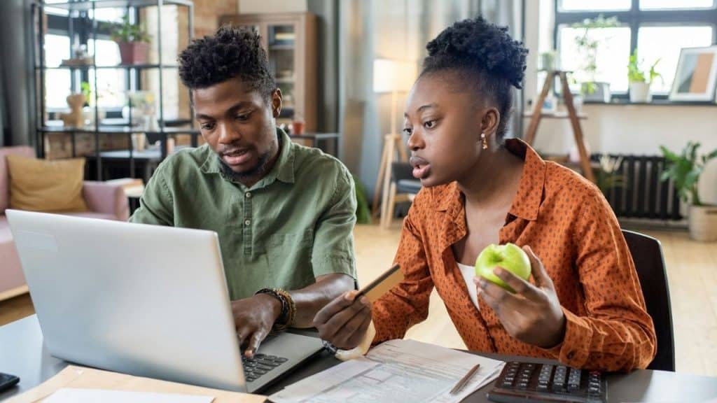 A man and woman sit at a desk with a laptop, calculator, and credit card.