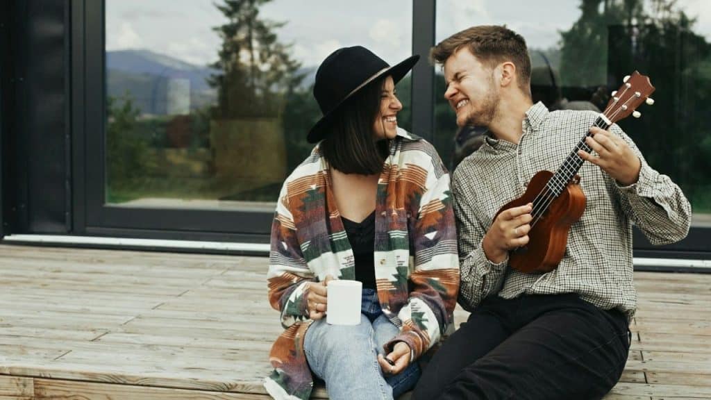 A woman with a mug and a man with a ukulele laugh together on a deck.