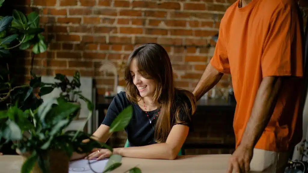 A woman smiling while writing at a table with someone standing beside her.