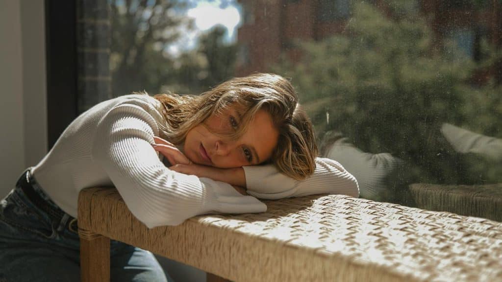 A person resting their head on folded arms beside a sunlit window.