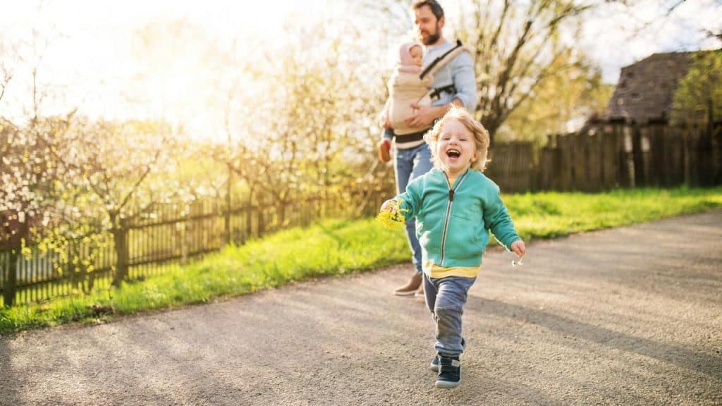 A toddler running with flowers while a parent carries a baby outdoors.