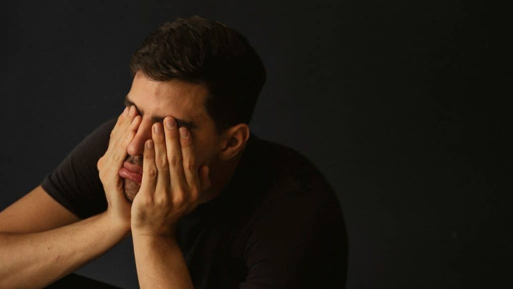 A man presses his hands against his face while looking downward against a dark background.