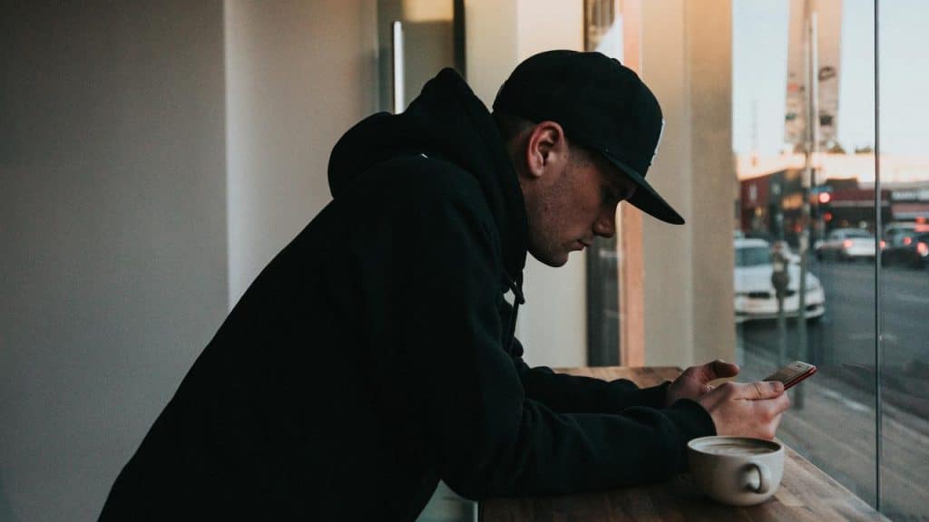 A man in a hoodie sitting by a window, checking his phone with a cup of coffee nearby.
