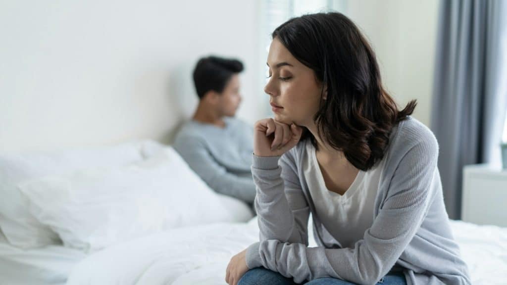 A woman sits on a bed looking down while a man sits blurred in the background.