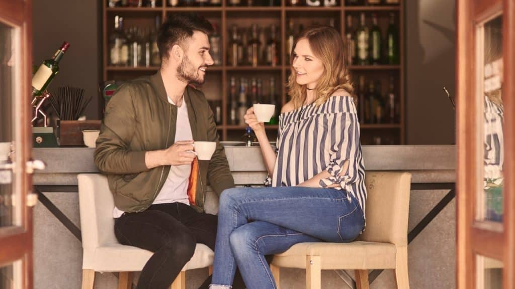 A man and woman sit at a bar counter holding white cups and looking at each other.