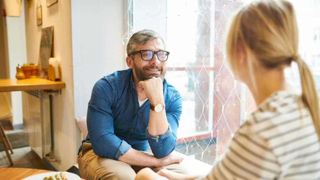 A man in a blue shirt sits by a window, smiling while looking at a woman.