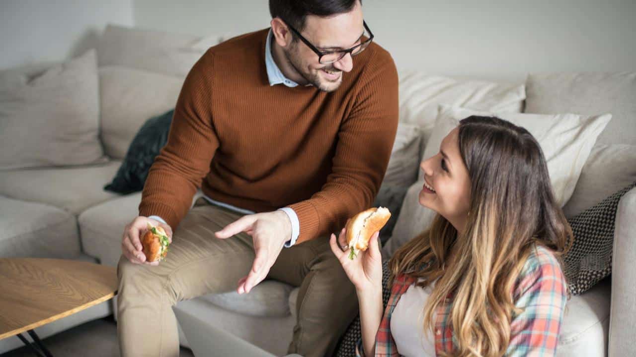 A smiling couple eats sandwiches while looking at a laptop in their living room.