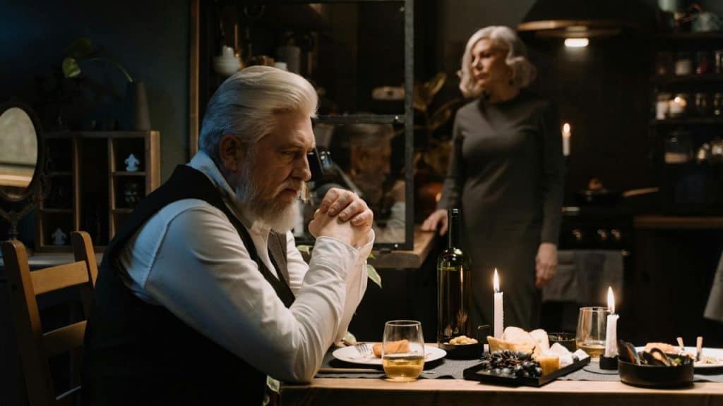 An older man with white hair sits at a candlelit table while a woman stands behind him.
