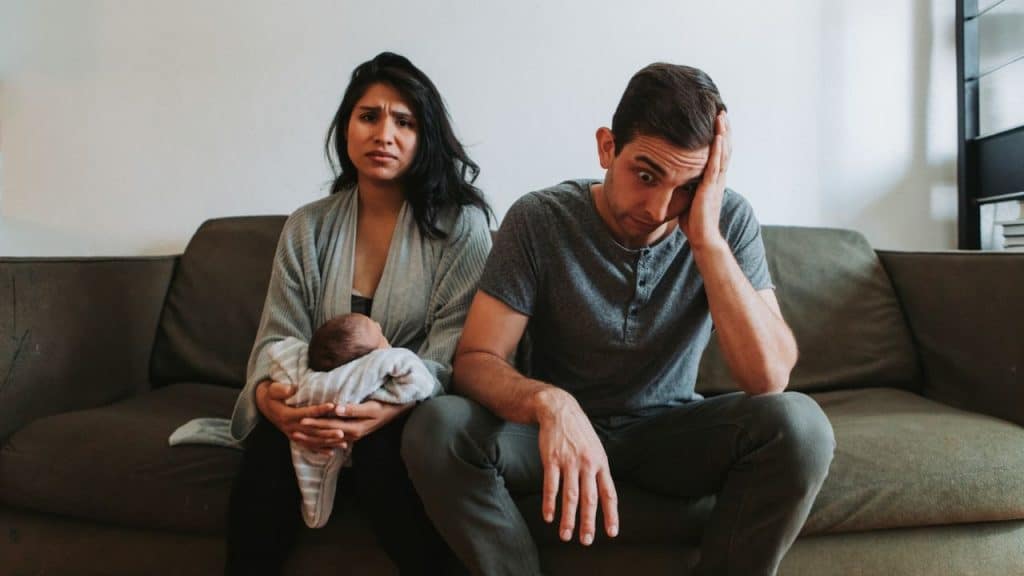 A man and woman sit on a sofa looking distressed while holding a small infant.