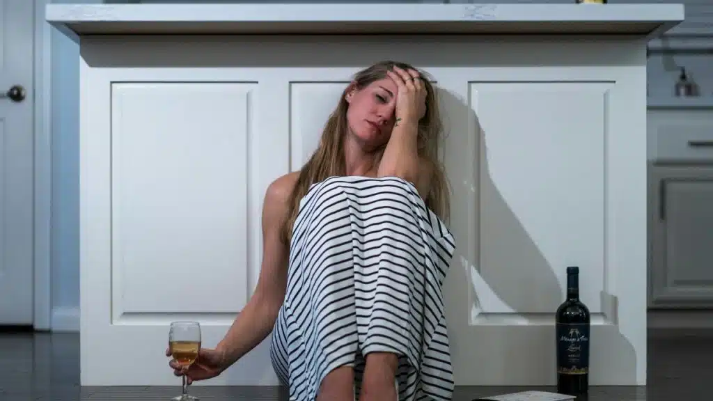 A woman in a striped dress sits on the floor holding a glass near a bottle.
