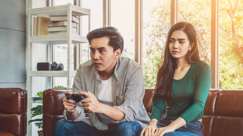 A man is playing a game on playstation and his annoyed wife is sitting with him.