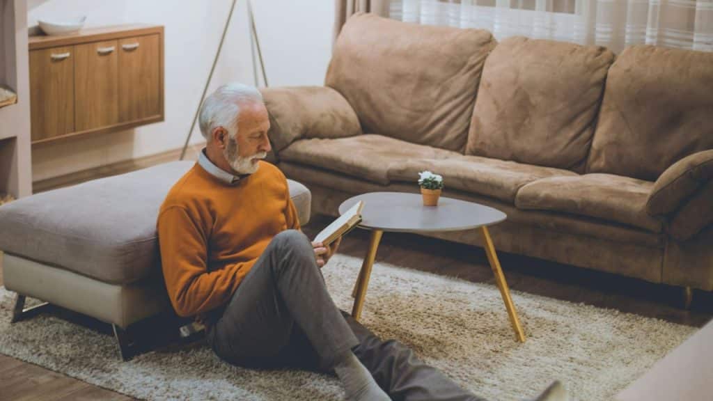 An older man with white hair sits on a rug while reading a small book.