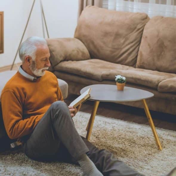 An older man with white hair sits on a rug while reading a small book.