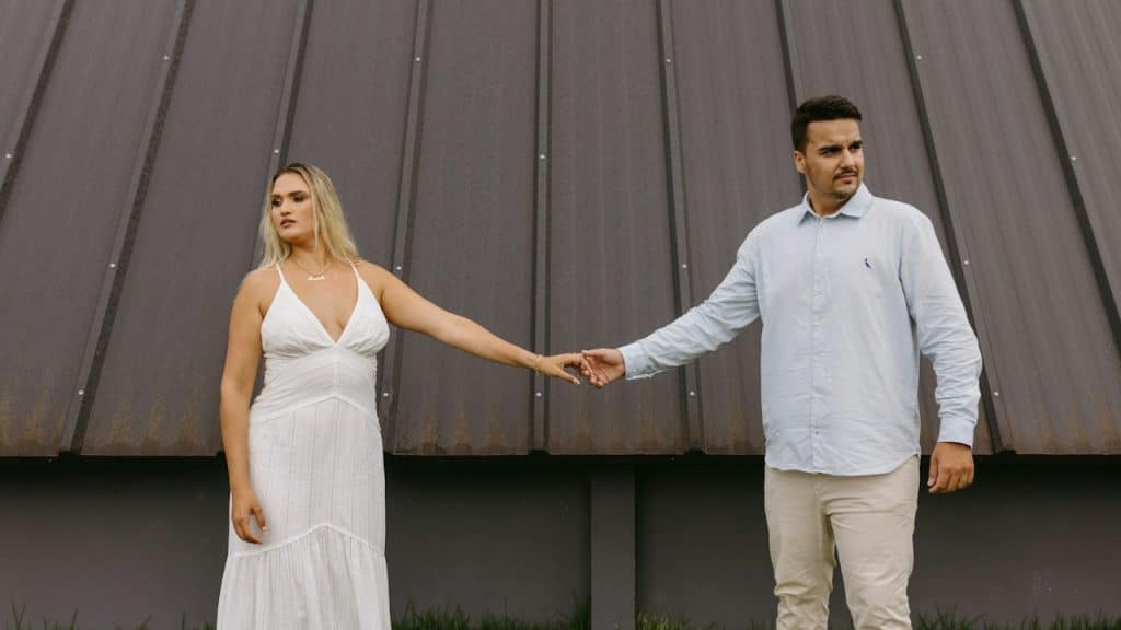 A couple standing apart while holding hands and looking in opposite directions.