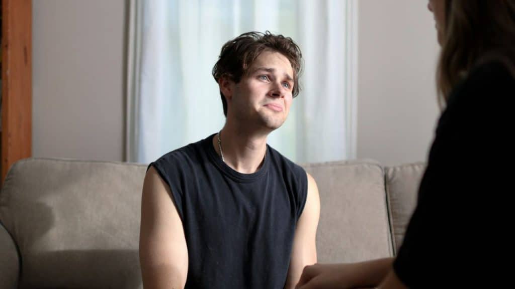A young man with a concerned expression sits on a couch and looks at someone off-camera.