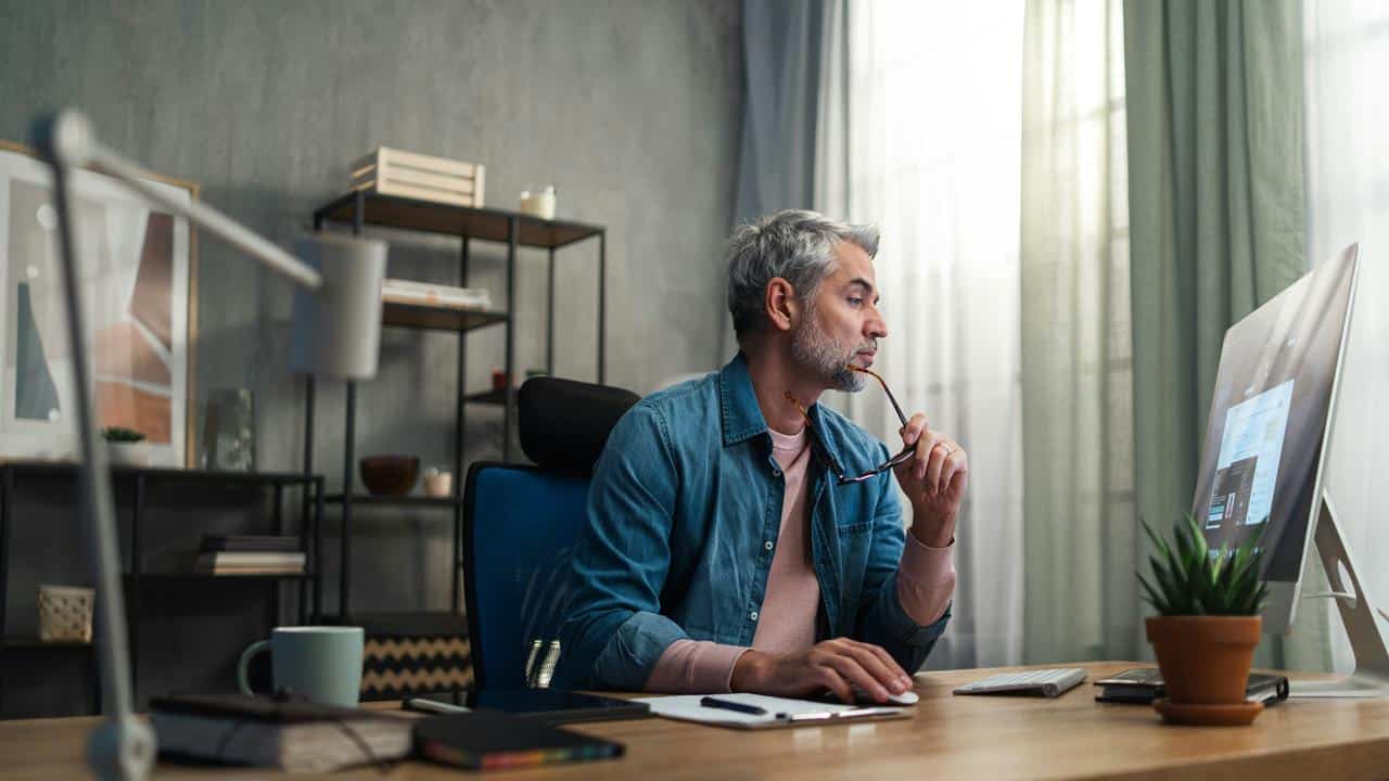 A middle-aged man working on a computer while holding eyeglasses on his hand.