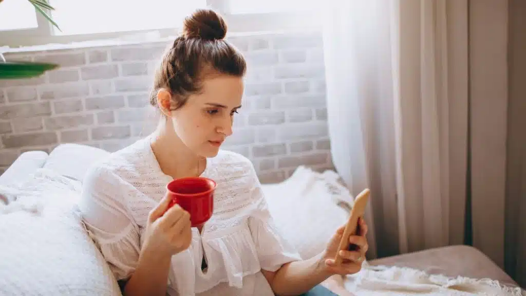 A woman with a hair bun holds a red mug and looks at her smartphone.