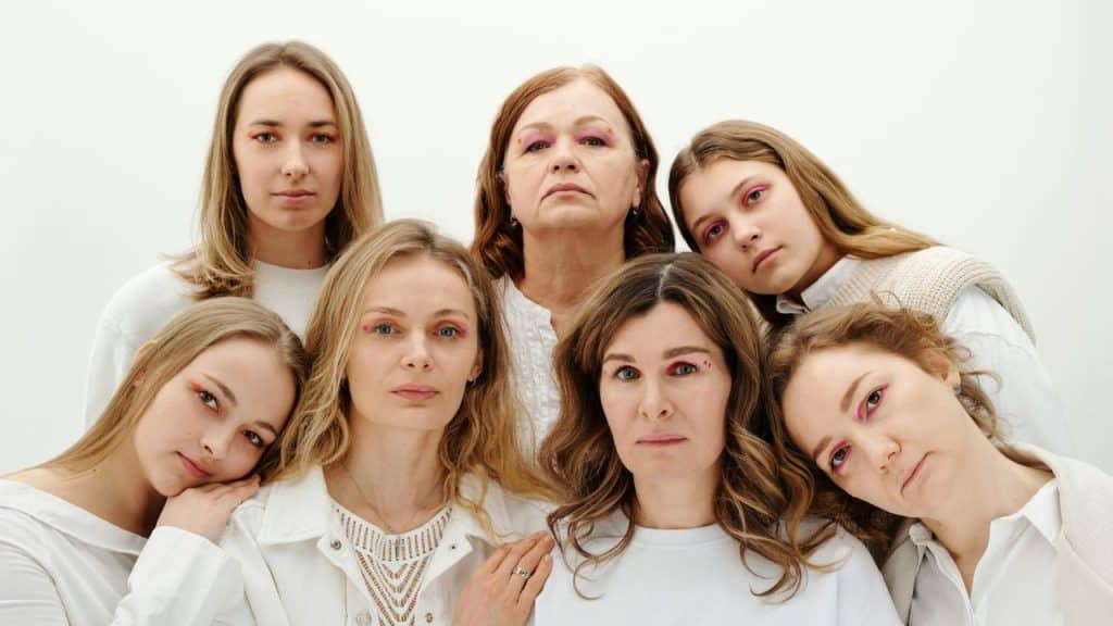 Seven women of various ages wearing white tops and pink eyeshadow pose for a group portrait.