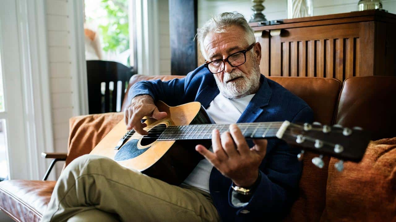 An older man with glasses playing an acoustic guitar on a couch.