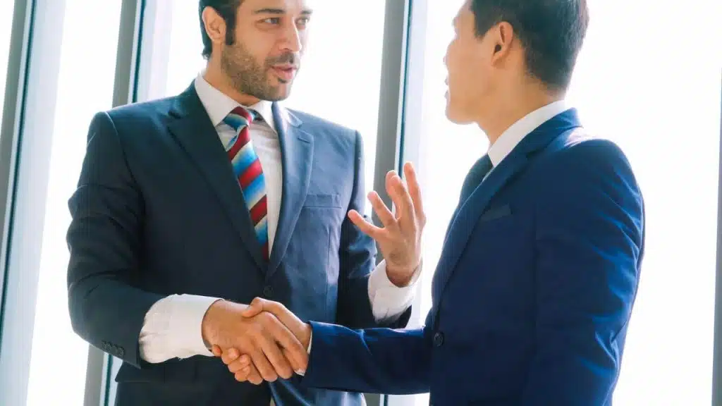 Two men in business suits shake hands while standing in front of a bright window.