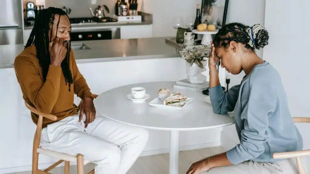 Two people sit at a table with food, both looking down with serious expressions.