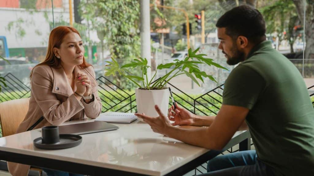A man and woman sit at a white table in a cafe near a plant.