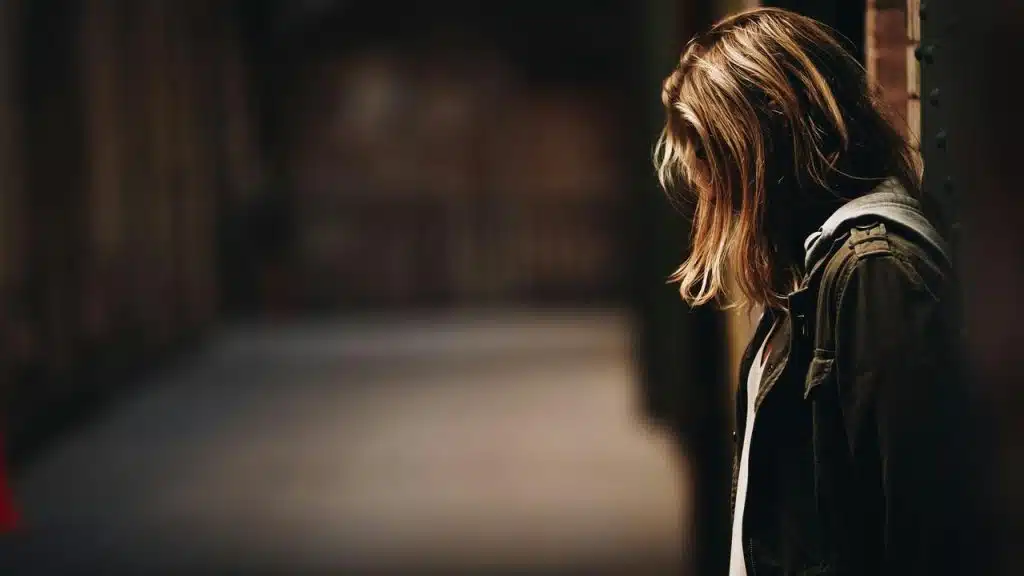 A person with shoulder-length hair looking down in a dimly lit alley.