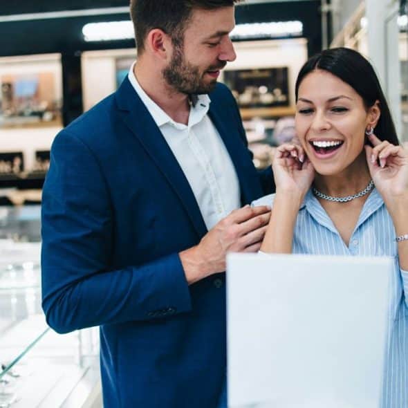 A smiling woman tries on silver earrings while a man in a blue blazer watches.