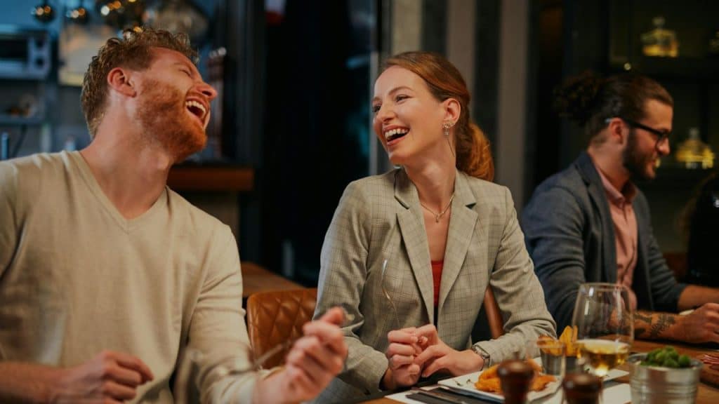 A man and a woman laugh together while sitting at a table in a crowded restaurant.
