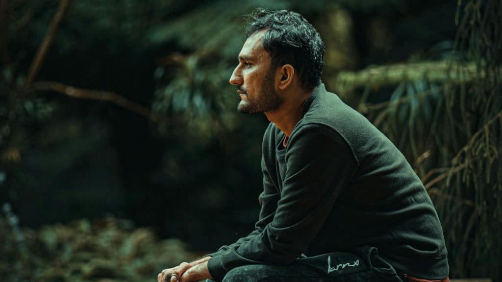 A man with a beard sits outdoors in profile view looking toward the dark woods.