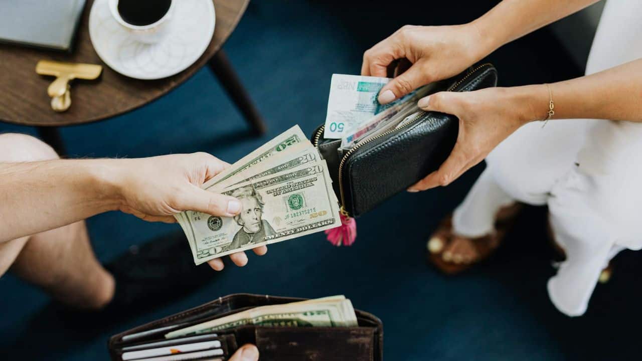 Two people hold cash and wallets over a blue surface next to a small wooden table.