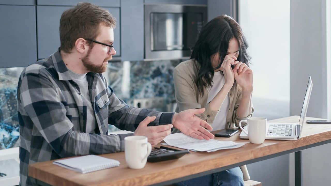 A man gestures toward paperwork at a table while a woman rubs her eyes nearby.