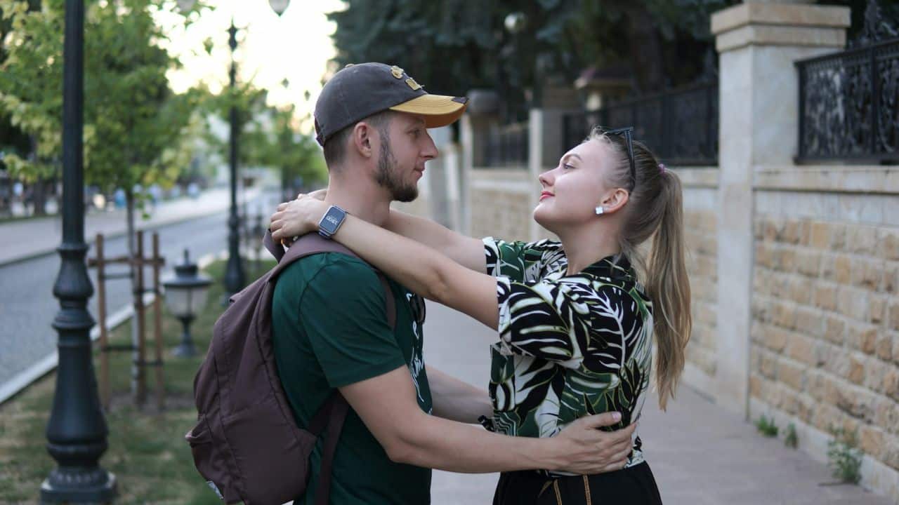 A woman with her arms around a man's neck looks up at him on a sidewalk.