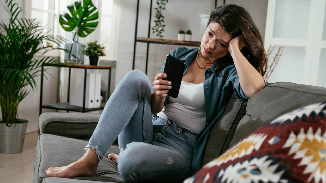 A woman sits on a grey sofa, leaning her head on her hand while looking at a smartphone.