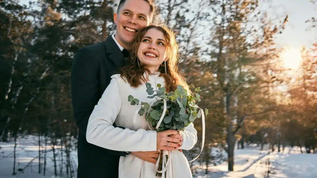 A smiling couple embracing outdoors in a snowy forest, with the woman holding a bouquet.